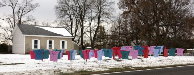 Image shows a memorial for victims of gun violence in Bucks County - foreground: t-shirts in different colors are stretched on stakes in the ground and placed in front of the meetinghouse - each t-shirt represents a certain number of gun violence victims. Snow is on the ground. Meetinghouse in background, with trees.
