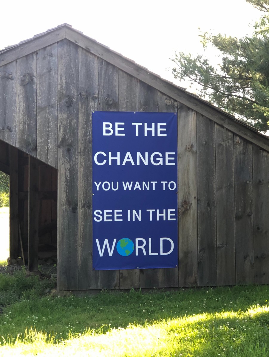 Photo shows a wooden structure on the meetinghouse grounds. The side of the structure in the photo has a large blue sign that says, in white writing, “Be the change you want to see in the world.” A picture of the Earth is the “O” in “world.” Grass and trees appear in the foreground and background.